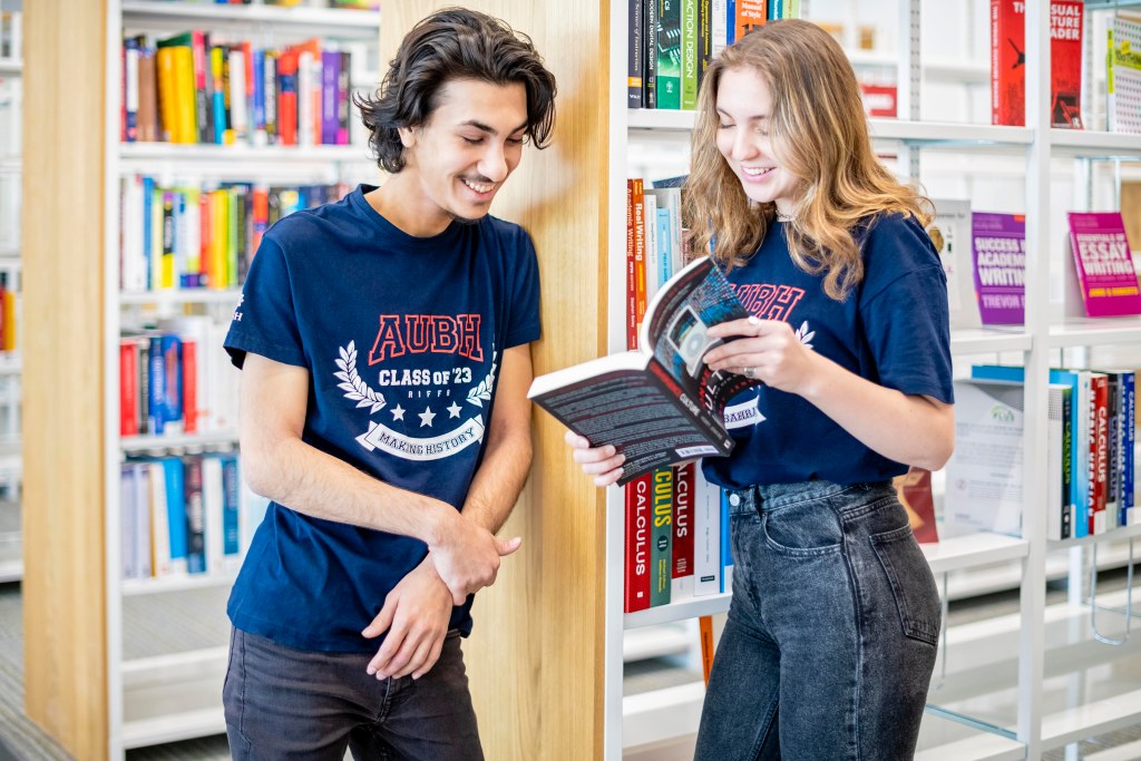 AUBH: two students sharing a book between library shelves, creative direction by Bashar Daas