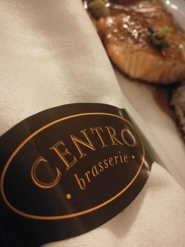 Centro Brasserie: the oval logo in gold on a dark brown napkin band, beside a plated dish, creative direction by Bashar Daas